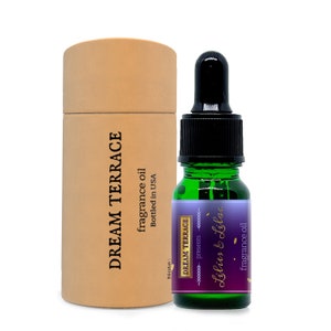 May include: A green glass bottle with a black dropper top and a purple and gold label that reads "DREAM TERRACE presents Lilies & Lilac fragrance oil". The bottle is next to a brown cardboard tube with the text "DREAM TERRACE fragrance oil Bottled in USA" printed on it.