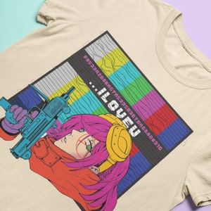 May include: A beige t-shirt with a graphic print of a woman with pink hair holding a gun. The graphic is set against a background of colorful, pixelated squares. The text "...ILOVEU" is printed on the graphic.