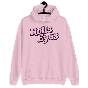 Puede incluir: Sudadera con capucha rosa con un gr&aacute;fico negro que dice "Rolls Eyes" en una fuente retro.