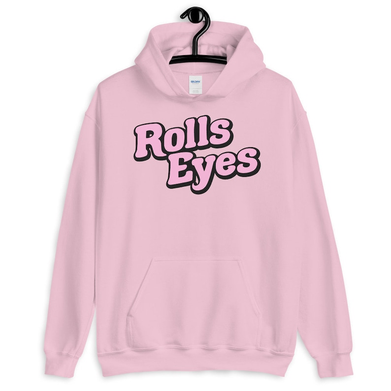 Puede incluir: Sudadera con capucha rosa con un gr&aacute;fico negro que dice "Rolls Eyes" en una fuente retro.