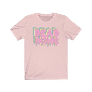May include: A light pink t-shirt with the words "DEAD INSIDE" in a dripping, layered design. The text is in pink, yellow, and blue. The shirt has short sleeves and a crew neck.