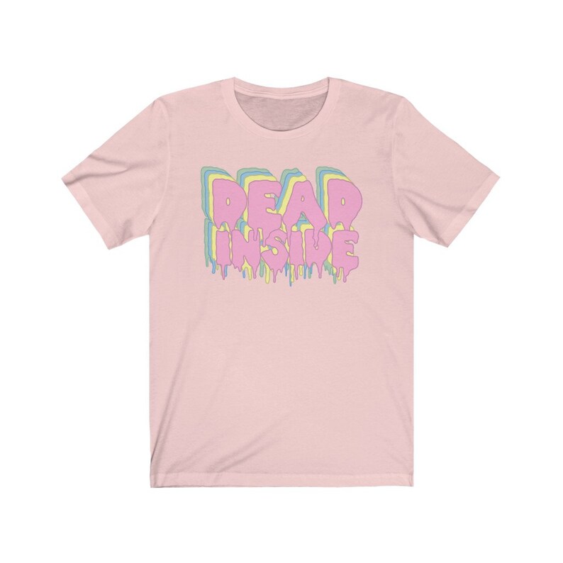 May include: A light pink t-shirt with the words "DEAD INSIDE" in a dripping, layered design. The text is in pink, yellow, and blue. The shirt has short sleeves and a crew neck.