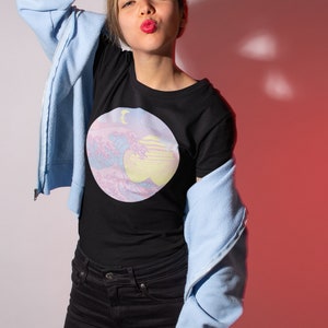 May include: A woman wearing a black t-shirt with a pastel pink and blue graphic of a wave and a crescent moon. She is also wearing a light blue zip-up hoodie and black jeans.