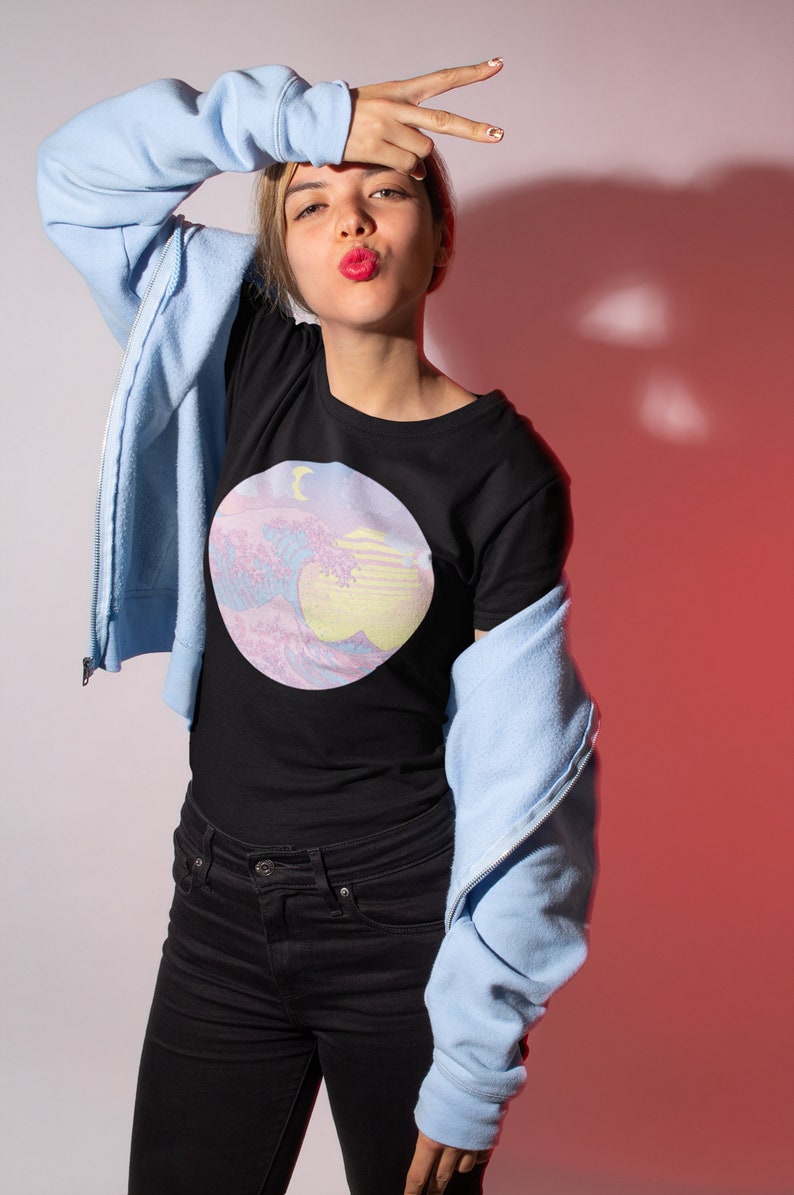 May include: A woman wearing a black t-shirt with a pastel pink and blue graphic of a wave and a crescent moon. She is also wearing a light blue zip-up hoodie and black jeans.
