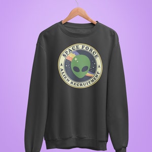Puede incluir: Una sudadera gris oscuro con un gráfico circular de un extraterrestre verde con un arcoíris y estrellas. El gráfico dice "Space Force Alien Recruitment".