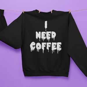 May include: Black sweatshirt with the text "I NEED COFFEE" in dripping white letters. The sweatshirt is hanging on a clothesline with wooden pegs against a purple background. A casual, comfortable apparel item.