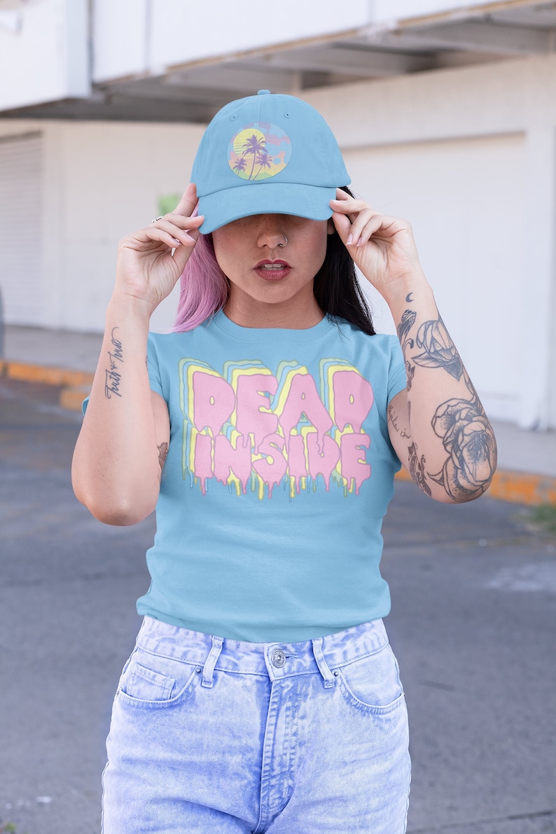 May include: Light blue baseball cap with a circular design featuring palm trees and a sunset. The model is wearing a light blue t-shirt with the text "DEAD INSIDE" in a dripping, layered design. The model is also wearing light blue jeans.