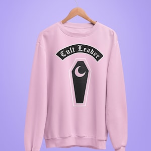 Puede incluir: Sudadera rosa con un gráfico en blanco y negro de un ataúd con una luna creciente y el texto "Cult Leader" en una fuente gótica.