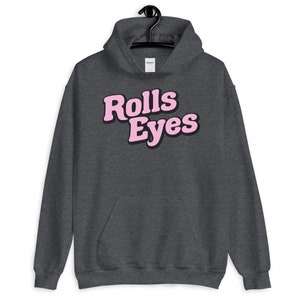 Puede incluir: Una sudadera con capucha gris oscuro con un gr&aacute;fico rosa que dice "Rolls Eyes" en una fuente retro.