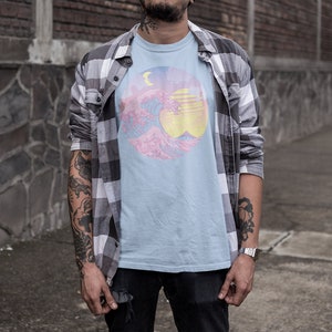 May include: A light blue t-shirt with a graphic print of a pink and blue wave with a yellow sun and a crescent moon. The t-shirt is worn by a person with tattoos on their arms.