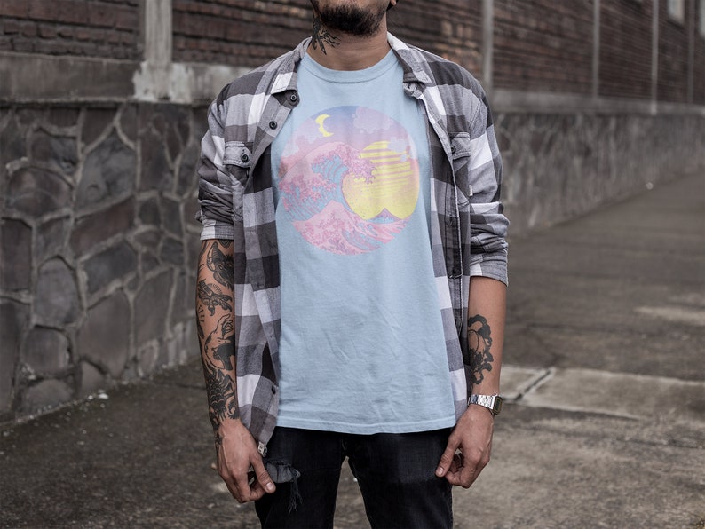 May include: A light blue t-shirt with a graphic print of a pink and blue wave with a yellow sun and a crescent moon. The t-shirt is worn by a person with tattoos on their arms.