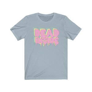May include: Light blue t-shirt with the text "DEAD INSIDE" in a dripping, layered design. The text is in pink, yellow, and blue. The shirt has short sleeves and a crew neck.