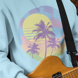 May include: A light blue hoodie with a graphic of a sunset and palm trees. The hoodie has a black strap across the chest.