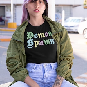 Op de afbeelding: Een persoon draagt een zwart T-shirt met de tekst "Demon Spawn" in een regenboogkleurige gradiënt, een groen jack, lichtblauwe jeans en een paarse baseballpet.