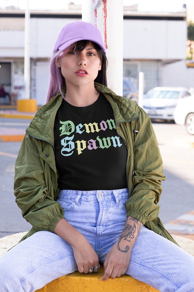 May include: A person wearing a black t-shirt with the text "Demon Spawn" in a rainbow gradient, a green jacket, light blue jeans, and a purple baseball cap.