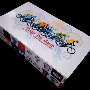 Pu&ograve; includere: Una scatola bianca con un'illustrazione colorata di ciclisti che corrono in bicicletta. La scatola &egrave; per un gioco da tavolo chiamato "Tour Du Jour: The World's Best Bicycle Racing Board Game".
