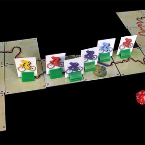 Pu&ograve; includere: Un gioco da tavolo con pedine di bicicletta rosse, gialle, blu, viola e verdi su una pista. Le pedine del gioco sono su una pista con una 'S' rossa per l'inizio. Un dado rosso a sei facce con numeri bianchi &egrave; sul tavolo.