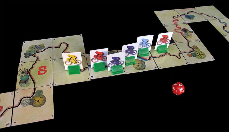 Pu&ograve; includere: Un gioco da tavolo con pedine di bicicletta rosse, gialle, blu, viola e verdi su una pista. Le pedine del gioco sono su una pista con una 'S' rossa per l'inizio. Un dado rosso a sei facce con numeri bianchi &egrave; sul tavolo.