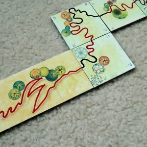 Pu&ograve; includere: Un tabellone da gioco con un percorso rosso e nero che si snoda attraverso una serie di tessere. Le tessere presentano illustrazioni di alberi e fiori. Il testo "Make the Magic Happen" &egrave; visibile sul tabellone.