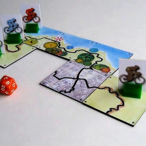 Pu&ograve; includere: Un gioco da tavolo con un tabellone colorato, dadi e pedine. Il tabellone presenta una strada tortuosa con alberi verdi e un oceano blu. Le pedine sono piccole figure di ciclisti.