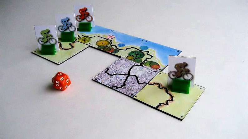 Pu&ograve; includere: Un gioco da tavolo con un tabellone colorato, dadi e pedine. Il tabellone presenta una strada tortuosa con alberi verdi e un oceano blu. Le pedine sono piccole figure di ciclisti.