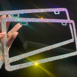May include: Two iridescent license plate frames encrusted with sparkling rhinestones. The frames are rectangular with rounded corners and feature mounting holes. The rhinestones reflect a rainbow of colors, creating a glamorous and eye-catching accessory.