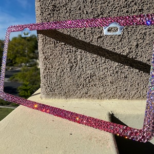May include: A pink and iridescent rhinestone license plate frame. The frame is made of metal and has a shiny finish.