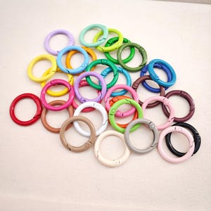 May include: A collection of circular metal spring gate rings in a variety of colors including red, yellow, blue, green, pink, purple, white, black, brown, and beige. Each ring features a hinged opening mechanism for attaching items.