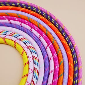 May include: A collection of braided ropes arranged in a semi-circular arc. The ropes feature a variety of vibrant colors including purple, orange, pink, light purple, white with multi-colored patterns, and bright yellow, all with a textured finish.