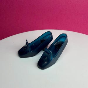 May include: A pair of dark blue miniature shoes with a low heel. The shoes have a classic loafer design with a rounded toe and a small decorative flap. The shoes are made of a translucent material.