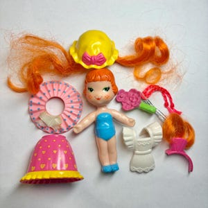 May include: A plastic doll with orange hair, freckles, and a blue swimsuit, accompanied by a yellow hat with a pink bow, a pink polka-dot skirt, a white ruffled dress, a pink flower accessory, and a pink hair clip with orange hair strands.