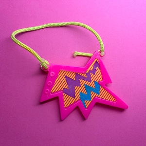 May include: A bright pink, star-shaped pendant necklace with a neon green cord. The pendant features a yellow and orange striped background with purple and blue zig-zag designs. The necklace has a retro, 80s or 90s aesthetic.