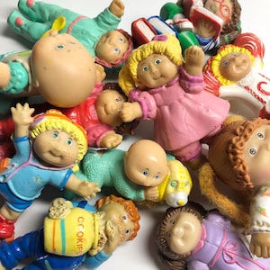 May include: A collection of Cabbage Patch Kids dolls in various outfits and poses. The dolls have different hair colors, skin tones, and facial expressions. Some dolls are wearing dresses, while others are in pajamas. One doll is holding a container that says "COOKIES".