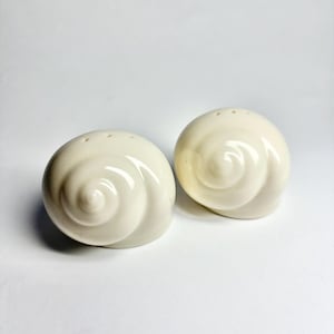 May include: A pair of off-white ceramic salt and pepper shakers shaped like spiral seashells. Each shaker features a smooth, glossy finish with a series of small holes at the top for dispensing seasoning, mimicking the natural form of a sea snail shell.