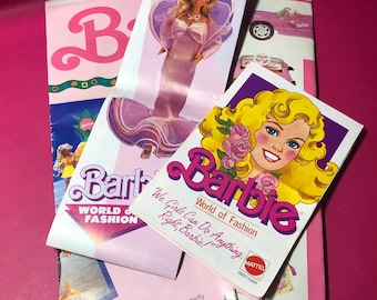 May include: A collection of Barbie doll packaging featuring a pink and white color scheme. The packaging includes the text "Barbie World of Fashion" and "We Girls Can Do Anything, Right Barbie!" along with a picture of a Barbie doll wearing a pink dress.