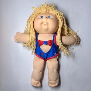 May include: A vintage Cabbage Patch Kid doll with blonde yarn hair and blue eyes. The doll is wearing a blue and red swimsuit with star accents and a red bow. The doll has a soft, plush body.