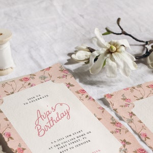 May include: A pink floral patterned invitation with the text "Join us to celebrate Ava's Birthday" and details for the event. The invitation is on a white surface with a spool of white ribbon and a white magnolia flower.