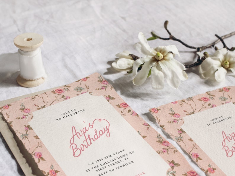 May include: A pink floral patterned invitation with the text "Join us to celebrate Ava's Birthday" and details for the event. The invitation is on a white surface with a spool of white ribbon and a white magnolia flower.