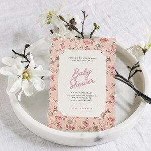 May include: A pink floral baby shower invitation with the text "Join us to celebrate Olivia Collin's Baby Shower" and details about the event. The invitation is on a white plate with a white magnolia flower.