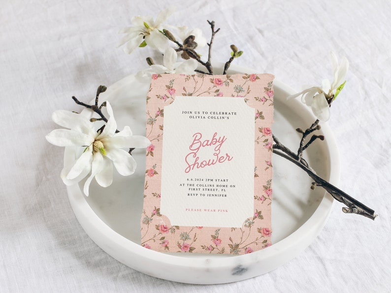 May include: A pink floral baby shower invitation with the text "Join us to celebrate Olivia Collin's Baby Shower" and details about the event. The invitation is on a white plate with a white magnolia flower.