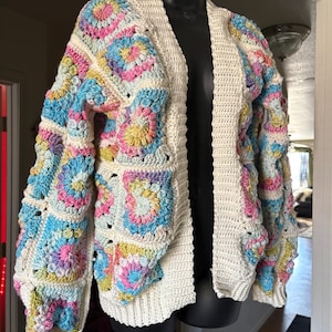 May include: A white crocheted cardigan with a colourful granny square pattern. The cardigan has long sleeves and an open front.