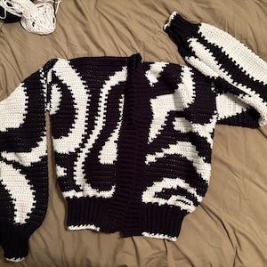May include: A handmade, open-front cardigan in a striking black and white pattern. The crochet design features bold, flowing white shapes against a black background. The sleeves are long and the cardigan is laid out on a neutral-toned surface.