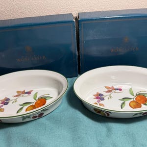 Può includere: Due pirofile ovali in ceramica bianca con bordo verde, ciascuna decorata all'interno con un motivo colorato di frutta e bacche. I piatti sono presentati davanti a due scatole di cartone blu scuro del marchio Royal Worcester.