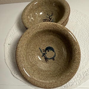 May include: Two round ceramic bowls with a speckled tan and brown finish. The front bowl features a hand-painted blue bird illustration in the center, while the bowl positioned behind it displays a painted pinecone design on its interior base.