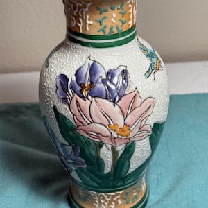 Può includere: Un vaso in ceramica con superficie bianca testurizzata, decorato con fiori dipinti a mano in rosa e viola, foglie verdi e una piccola farfalla blu. Il collo e la base presentano fasce arancioni e verdi, con un sottile bordo color oro sul colletto.