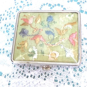 May include: A rectangular metal compact case featuring a light green fabric top panel embroidered with a multi-colored floral pattern in shades of pink, blue, yellow, and white. The case has a polished silver-tone metal frame and a small latch.
