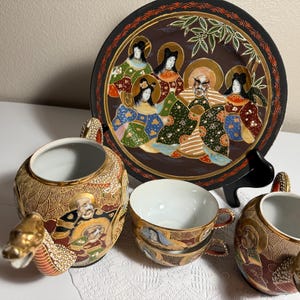 Könnte beinhalten: Ein Teeservice bestehend aus einer Teekanne, zwei Teetassen und einem Zierteller, verziert mit komplexen goldenen, schwarzen und bunten Mustern, die Figuren darstellen. Die Teekanne und ein kleineres Gefäß haben drachenförmige Ausgüsse.
