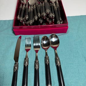 May include: A flatware set featuring a knife, two forks, and two spoons arranged in front of a red box containing additional utensils. Each piece has a polished silver-tone metal head and a black handle with ornate, embossed silver-tone metal accents.