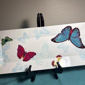 May include: A rectangular white ceramic serving platter featuring a pattern of butterflies. The design includes one large blue butterfly, one magenta butterfly, one small green butterfly, and several light blue butterfly outlines.