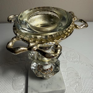 May include: A decorative object featuring a clear glass bowl with a gold-toned rim and two ornate gold-toned handles. The bowl sits atop a clear, faceted glass pedestal base, which is mounted on a square, white marble base with grey veining.
